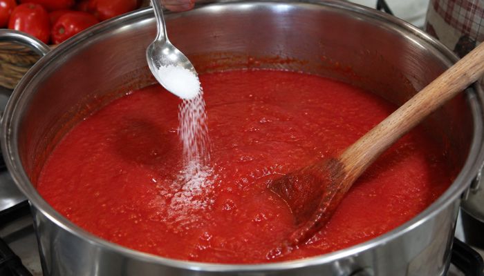 Un cucchiaio aggiunge sale a una pentola di passata di pomodoro mentre un cucchiaio di legno viene utilizzato per mescolare.
